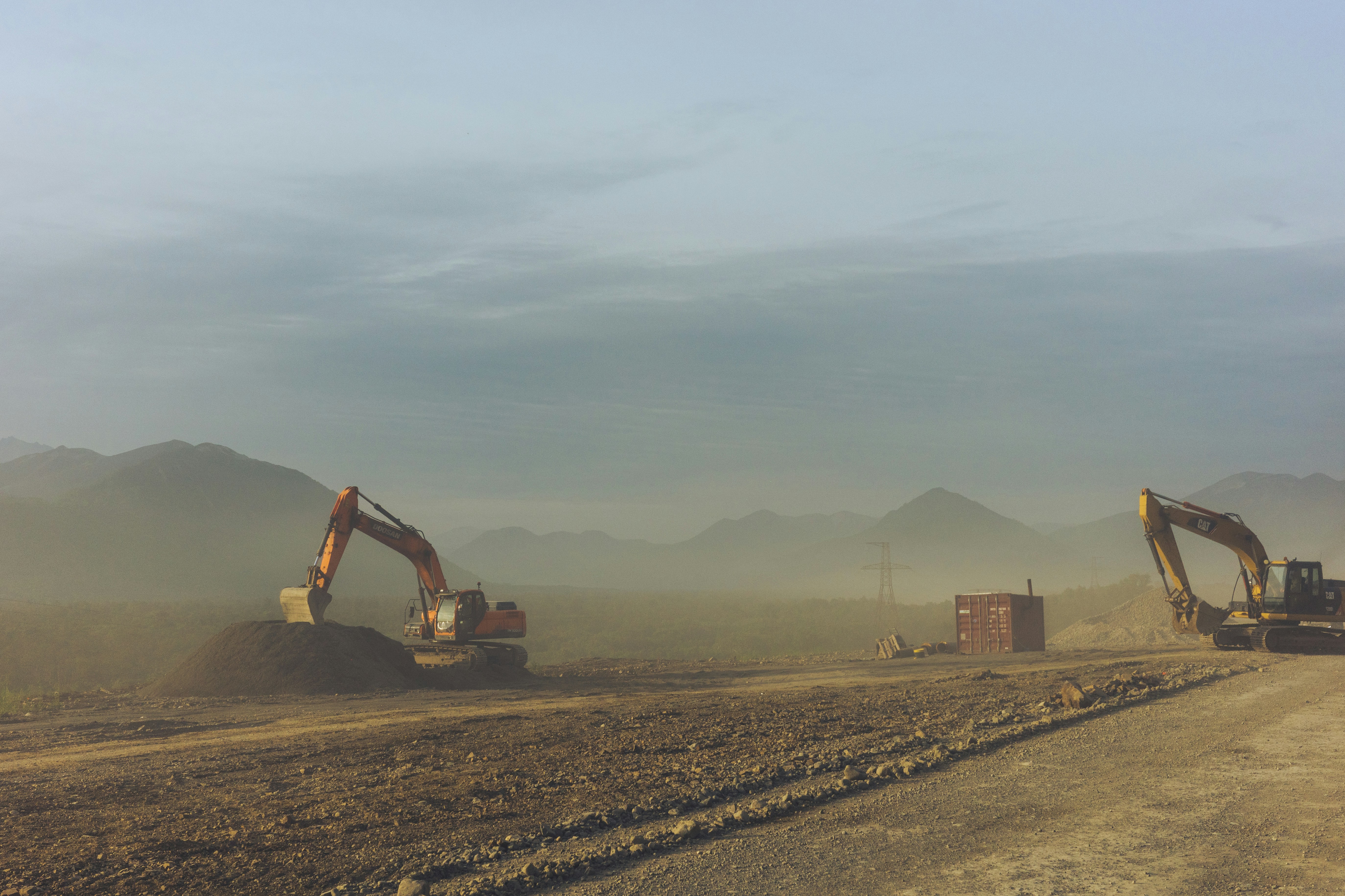 Two crawler excavators performing earthmoving tasks in a dusty construction site with mountains in the background, illustrating heavy machinery fleet operations.